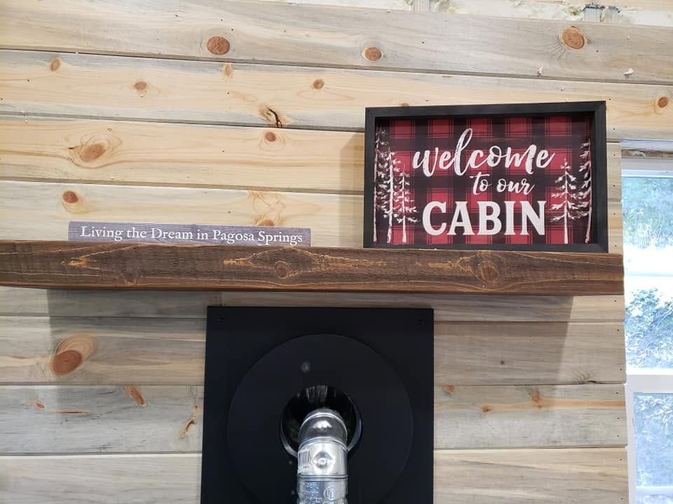 Welcome to our cabin plaid sign on a pine wall above a reclaimed wood mantel